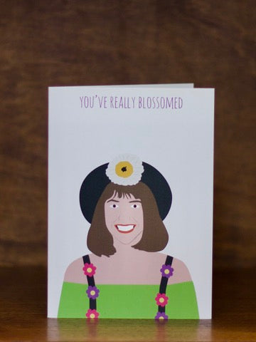 All occasions greeting card with illustration of a woman with a flower in her hair and text 'You've really blossomed' on a wooden surface.