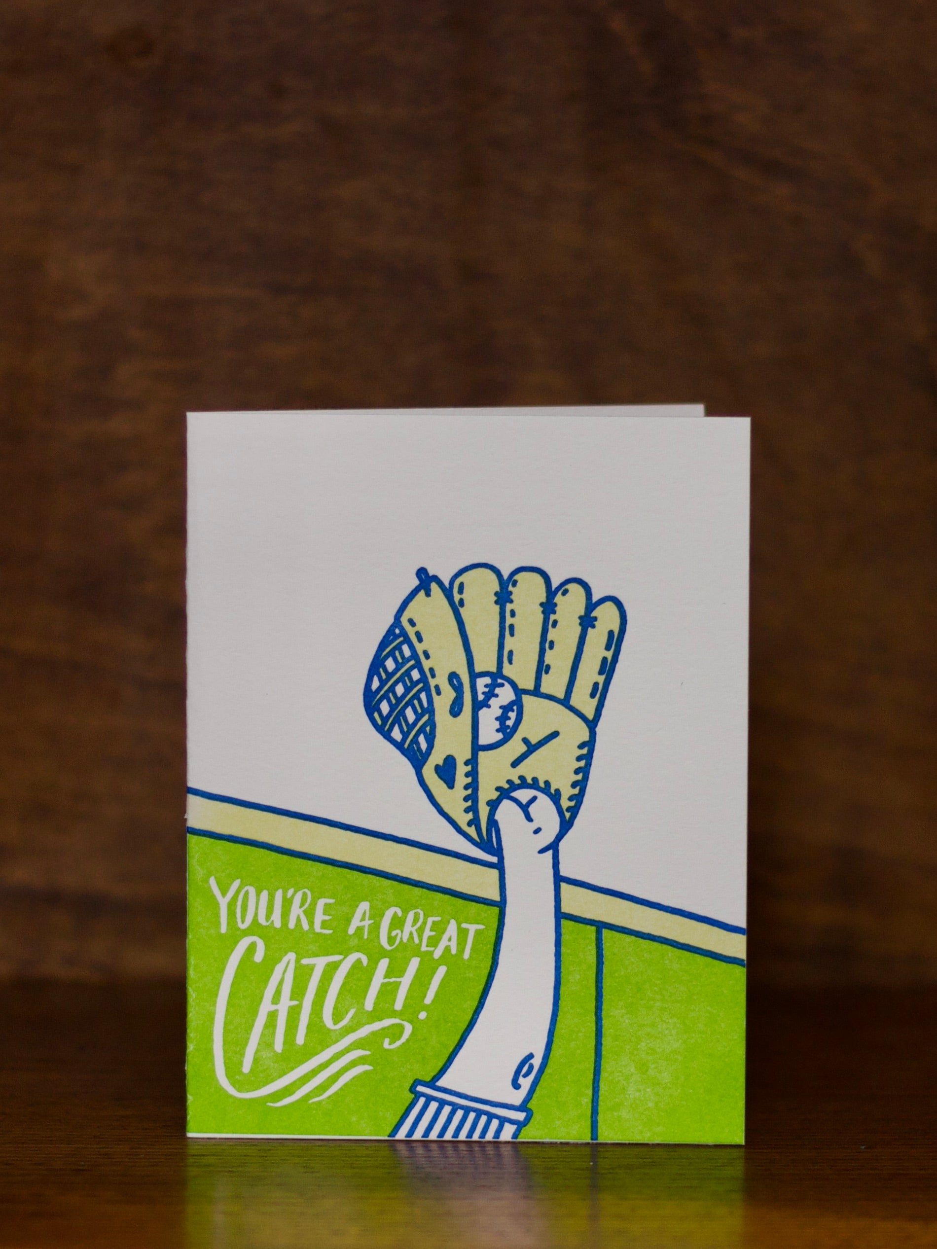 Greeting card with a hand holding a baseball glove and text 'You're a great catch!' on a wooden surface by Bench Pressed Greeting Cards