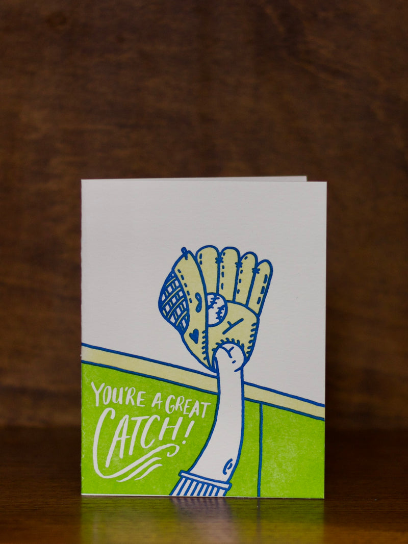 Greeting card with a hand holding a baseball glove and text 'You're a great catch!' on a wooden surface by Bench Pressed Greeting Cards