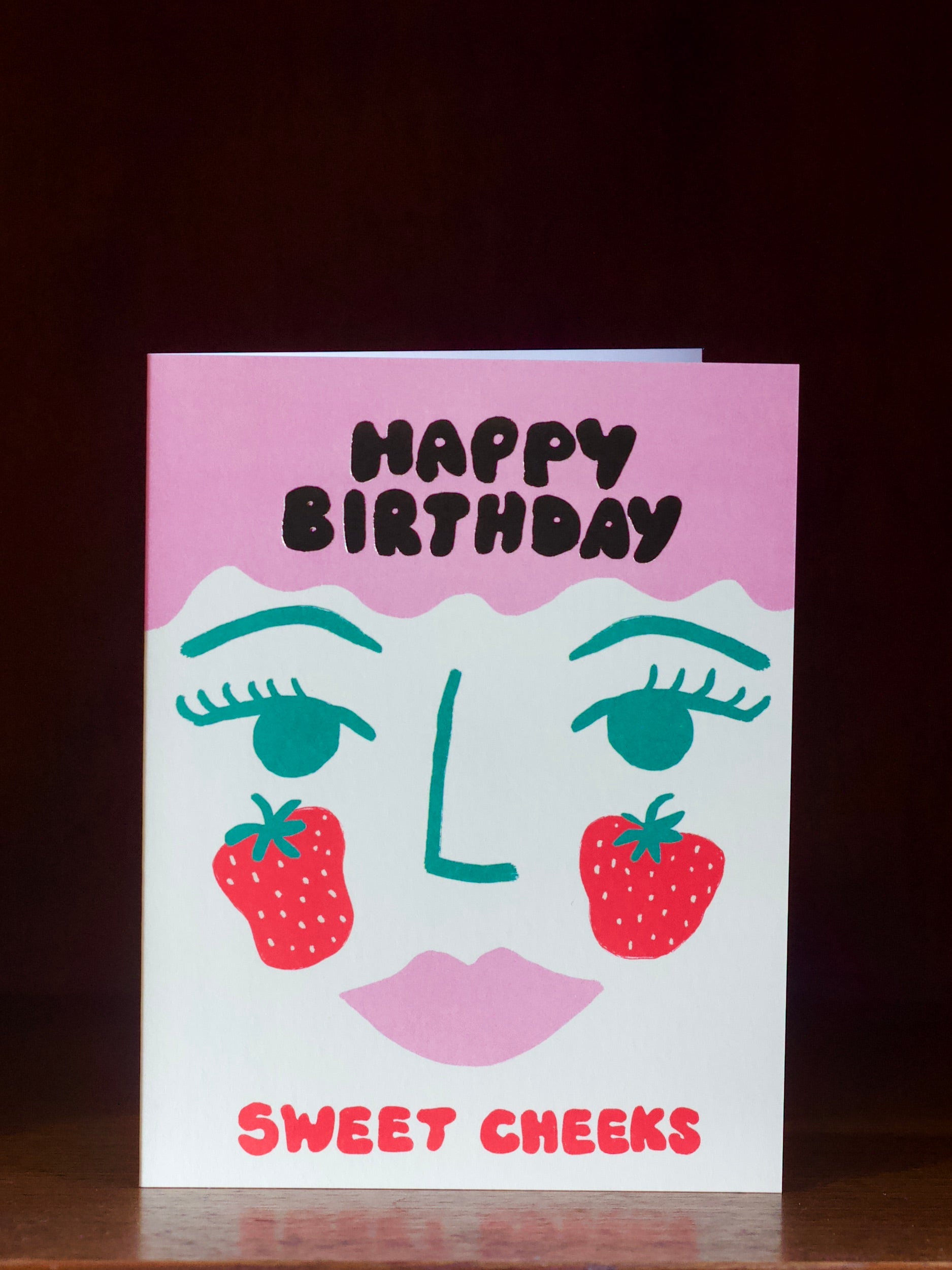 Birthday card with face design, strawberries, and 'Happy Birthday Sweet Cheeks' text on a dark background