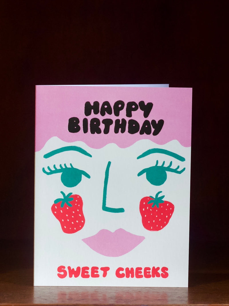 Birthday card with face design, strawberries, and 'Happy Birthday Sweet Cheeks' text on a dark background