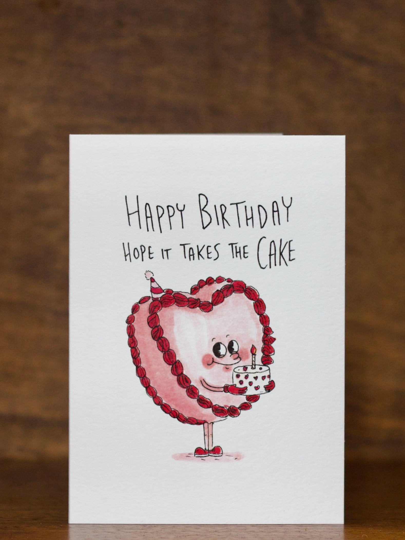 Birthday greeting  card by Well Drawn with cartoon cake character and 'Happy Birthday, hope it takes the cake' text on a wooden background