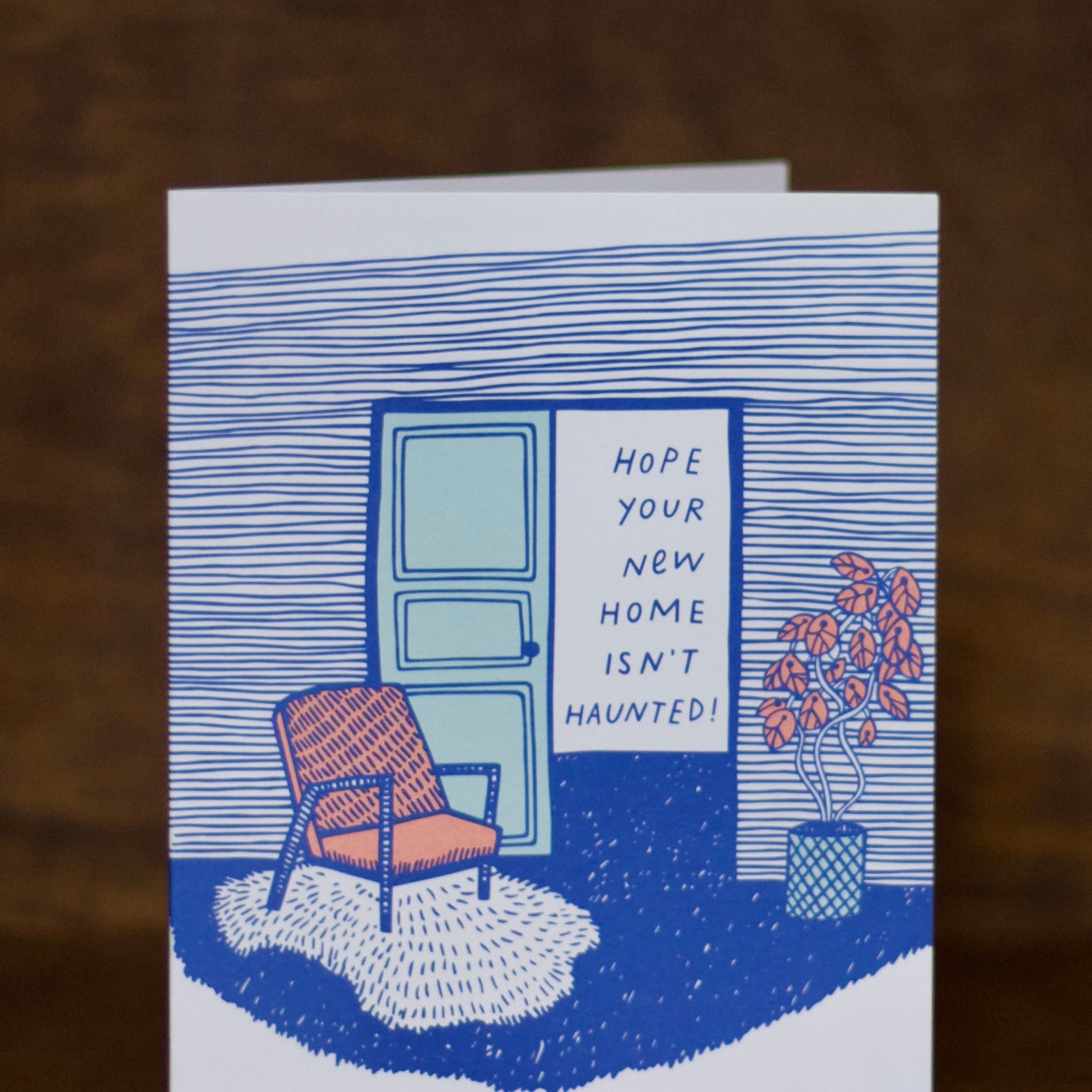 New home greeting card with a blue door, chair, and plant illustration on a brown background