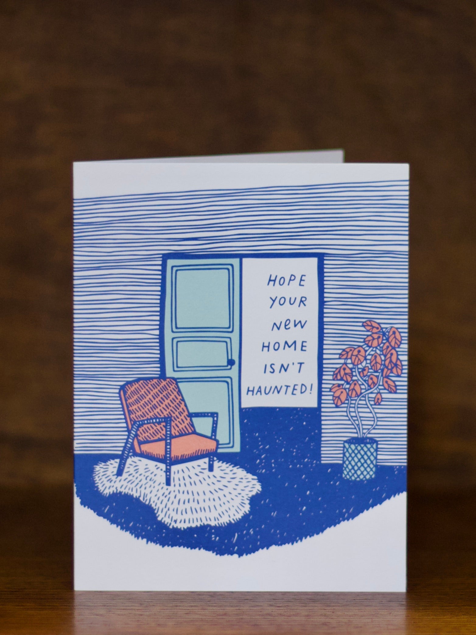 New home greeting card with a blue door, chair, and plant illustration on a brown background