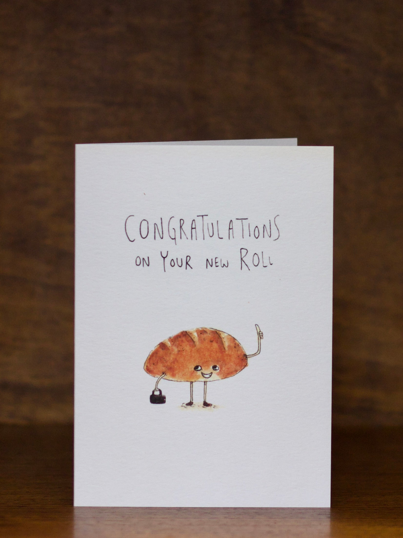 Congratulations greeting card with a cartoon croissant character and text 'Congratulations on your new roll' on a brown background
