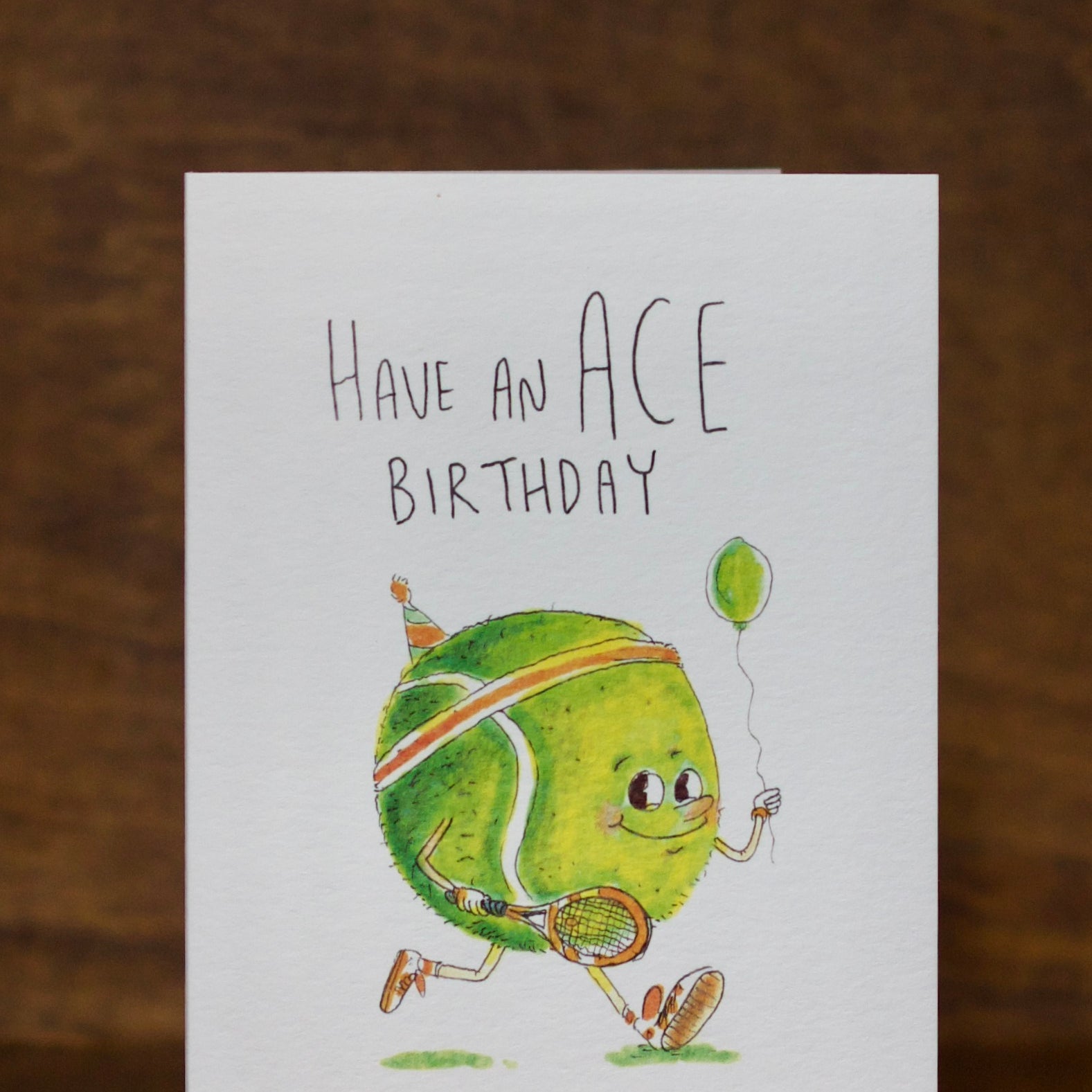 Birthday Greeting card by Well Drawn with a tennis ball character holding a balloon, text 'Have an ACE Birthday', on a wooden surface.