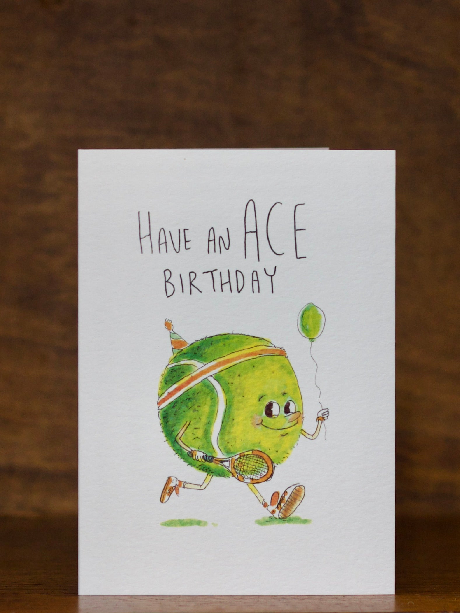 Birthday Greeting card by Well Drawn with a tennis ball character holding a balloon, text 'Have an ACE Birthday', on a wooden surface.