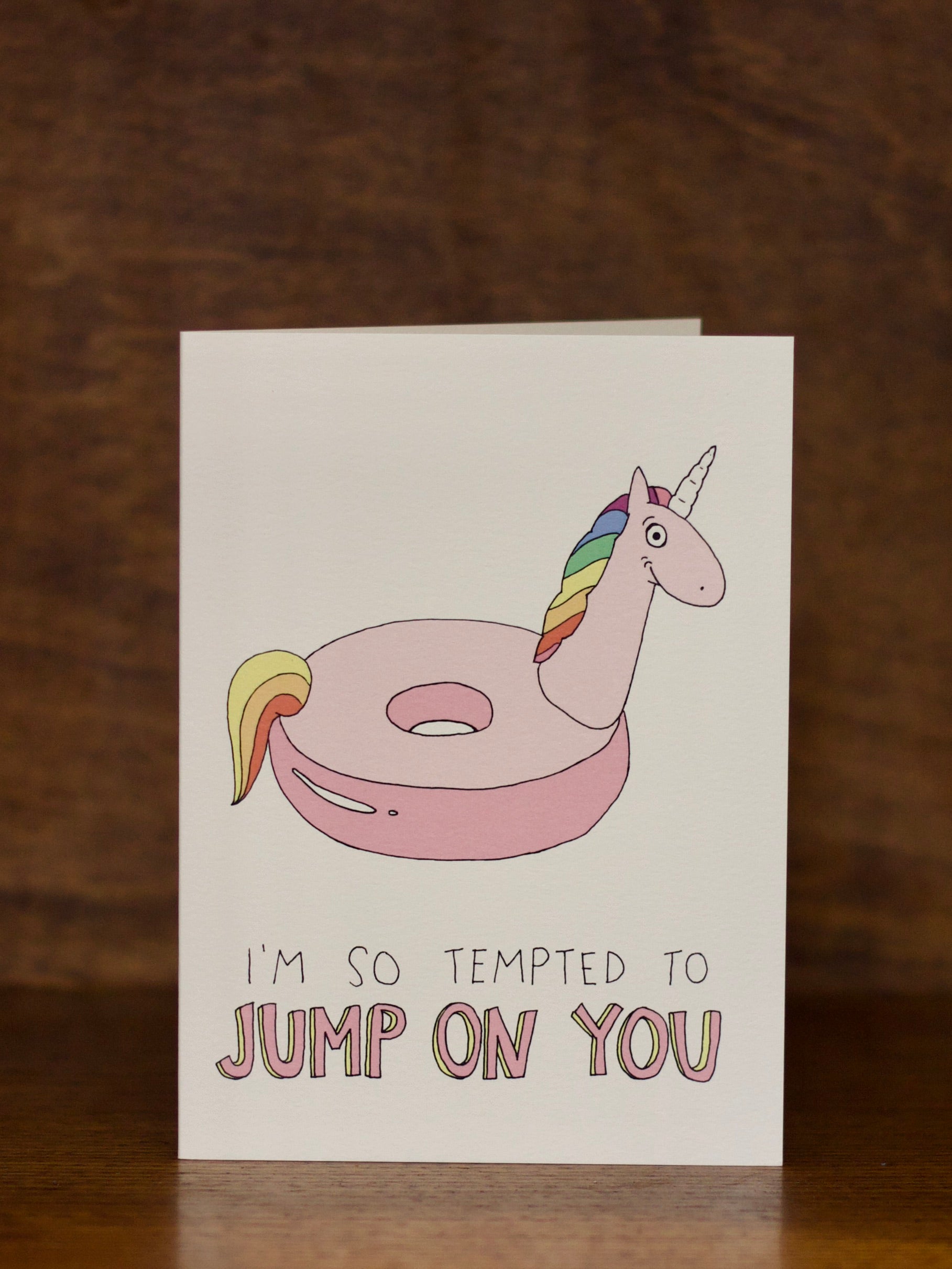 Love & Friendship greeting card with a unicorn on an inflatable ring and text 'I'm so tempted to jump on you' on a wooden surface.