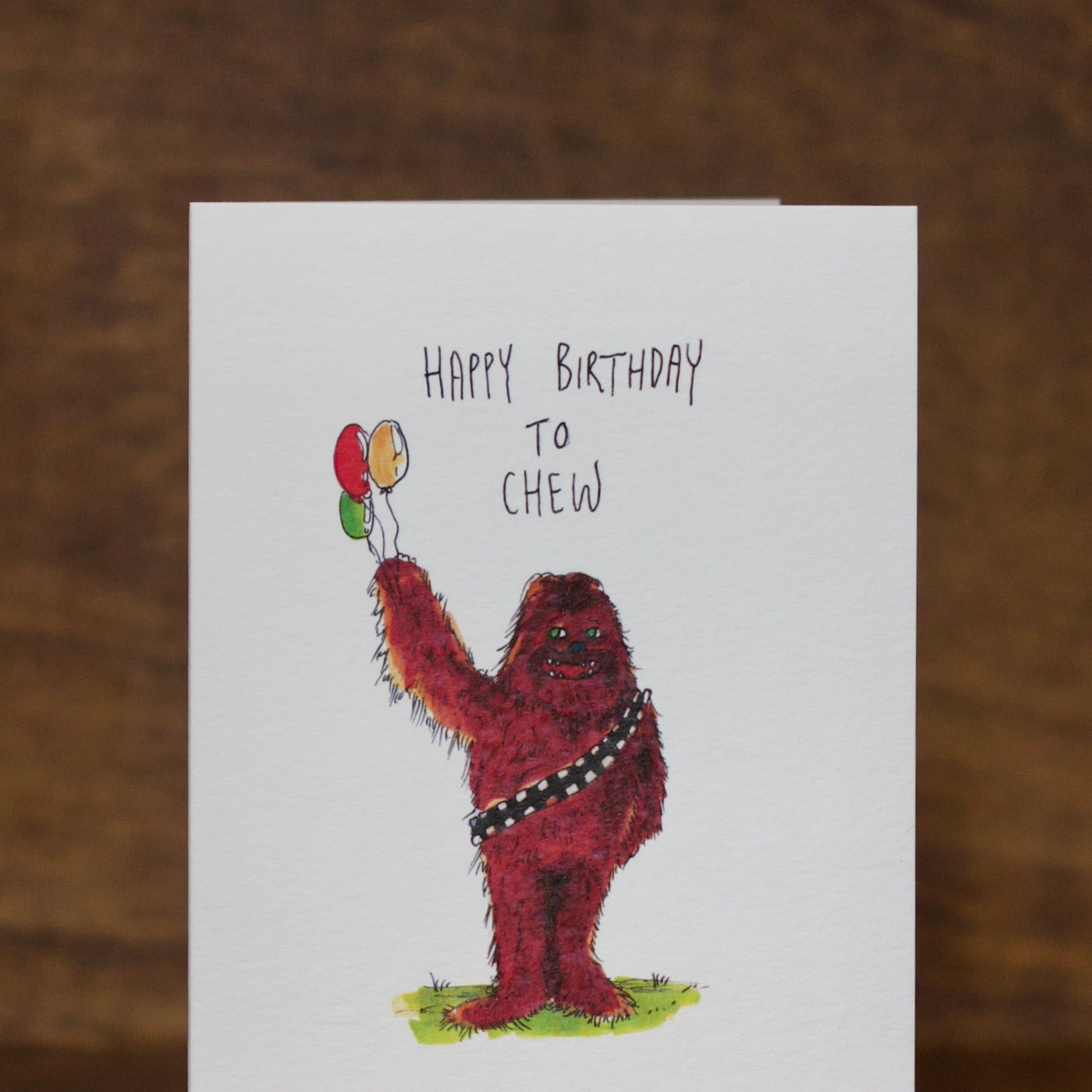 Birthday greeting card  by Well Drawn with a character holding balloons and text 'Happy Birthday to Chew' on a wooden surface.