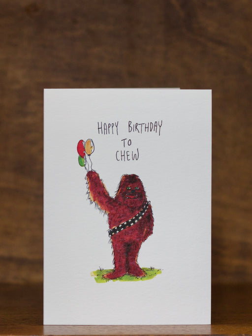 Birthday greeting card  by Well Drawn with a character holding balloons and text 'Happy Birthday to Chew' on a wooden surface.