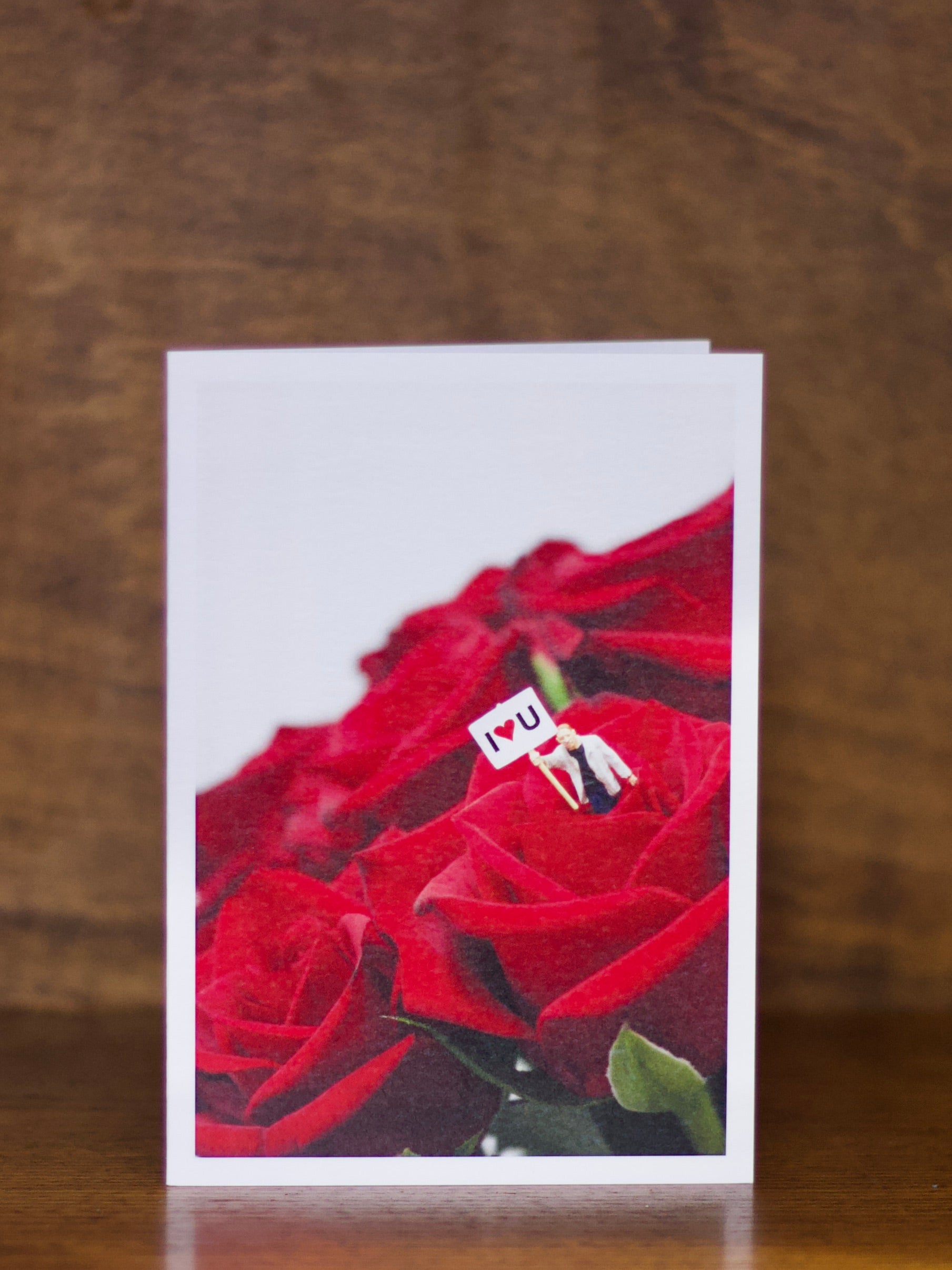 Love & Friendship greeting card with red roses and a small figure holding a 'I U' sign on a wooden surface.
