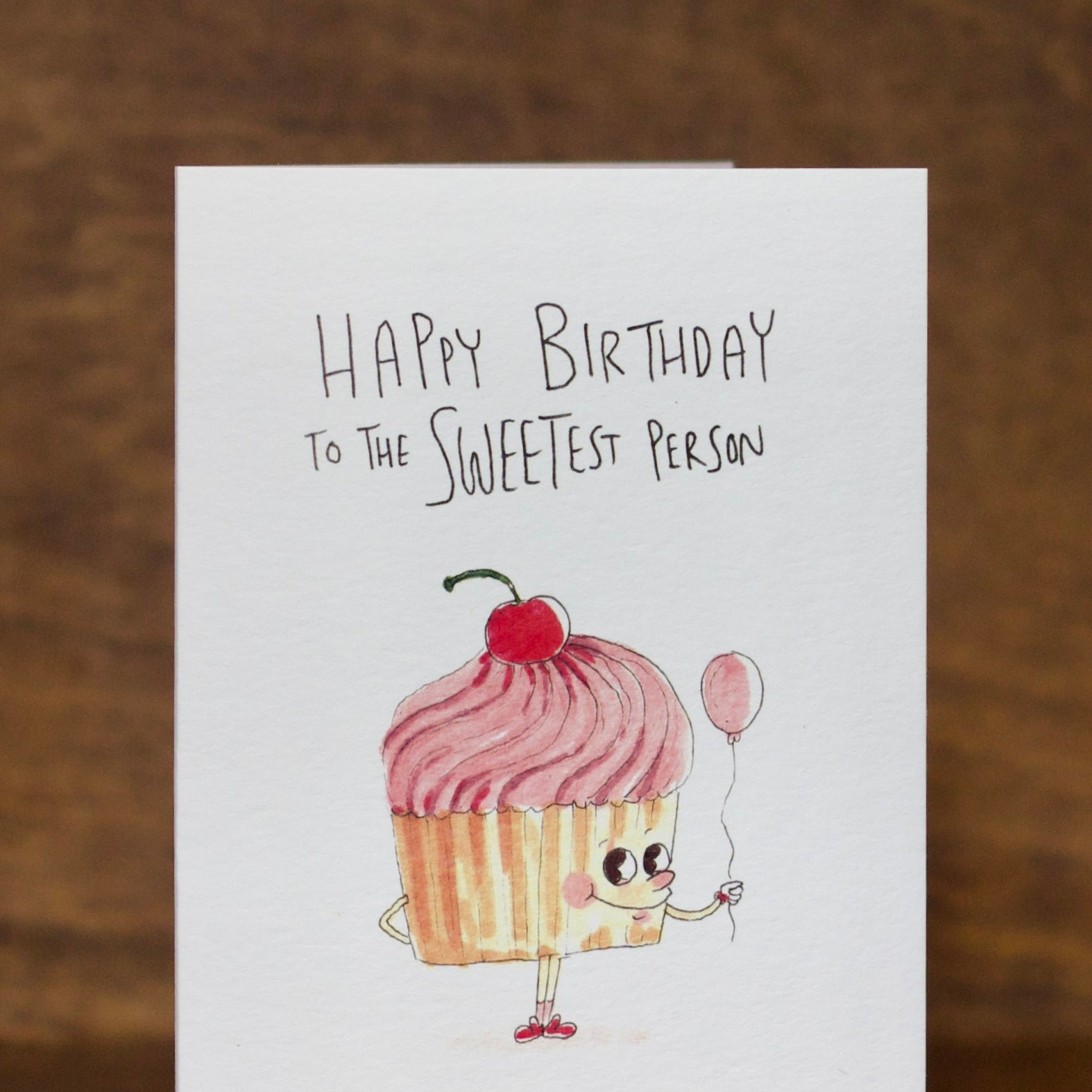 Birthday greeting card by Well Drawn with a cartoon cupcake character holding a balloon on a wooden surface.