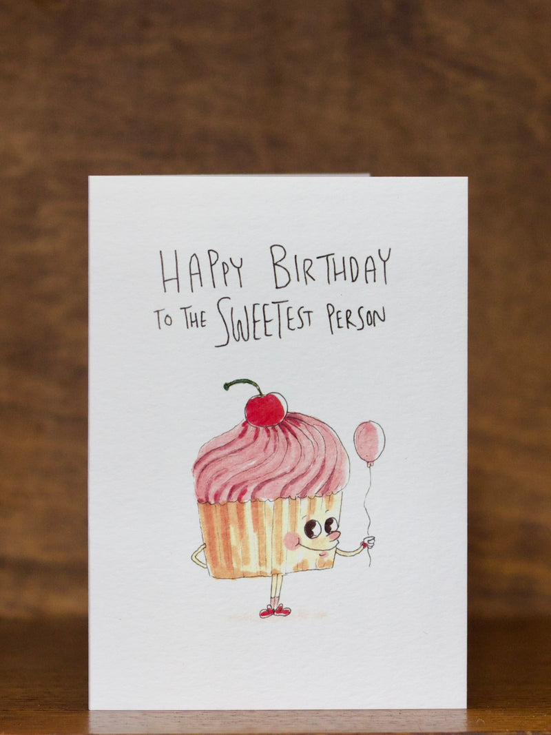 Birthday greeting card by Well Drawn with a cartoon cupcake character holding a balloon on a wooden surface.