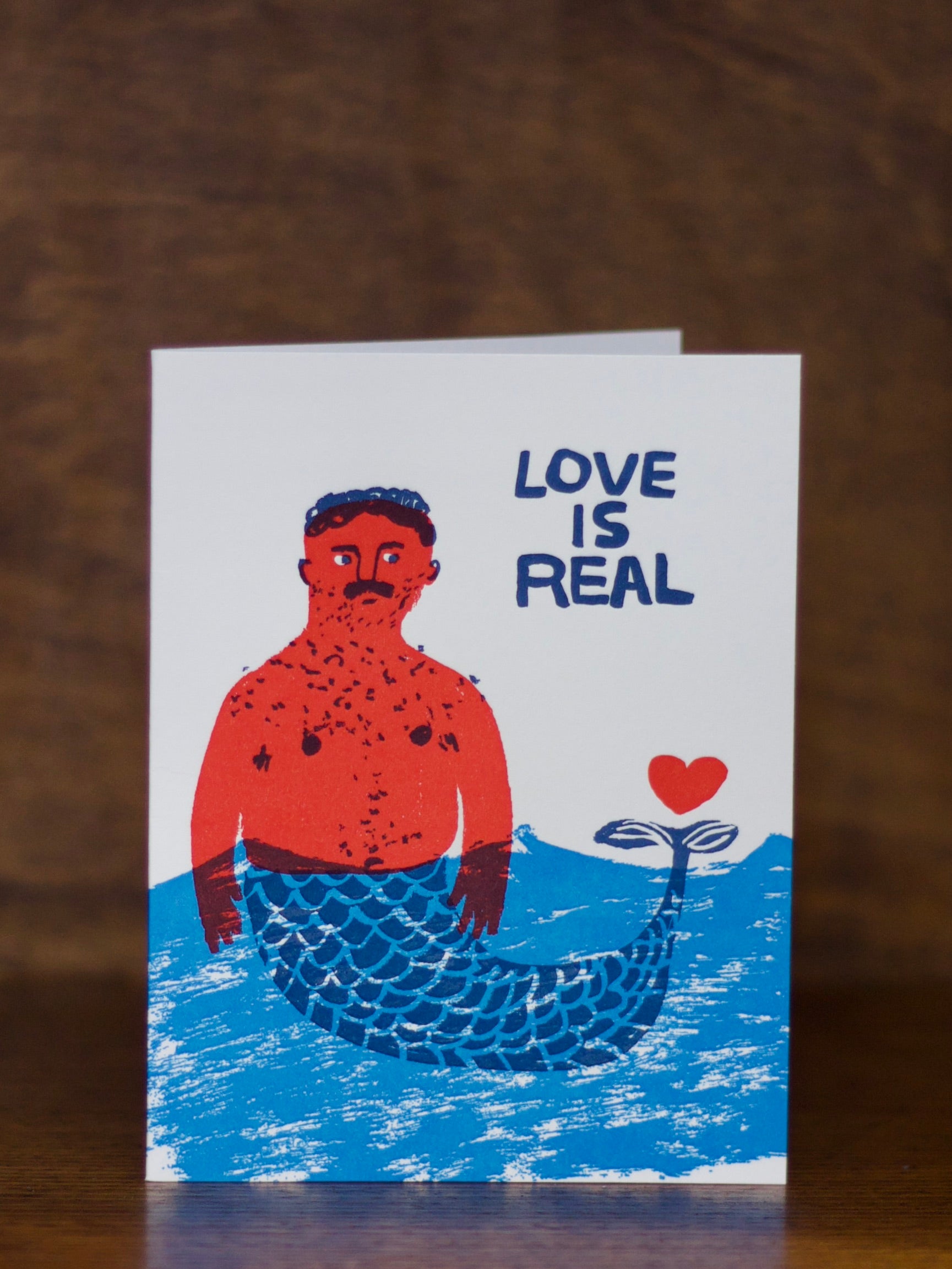 Love & Friendship greeting card with a mermaid illustration and 'Love is Real' text on a wooden surface