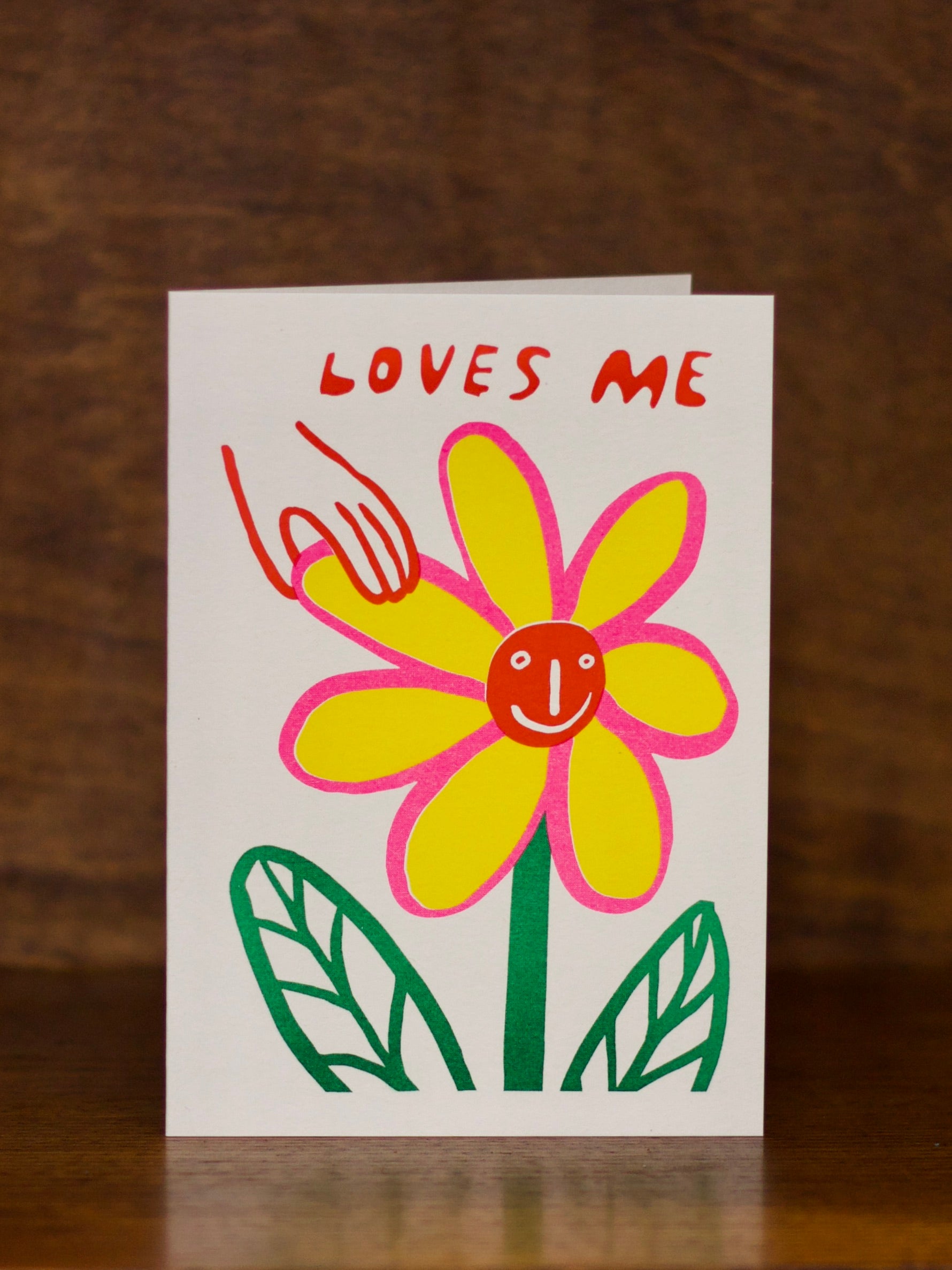 Love & Friendship greeting card with a yellow flower and text 'LOVES ME' on a wooden surface