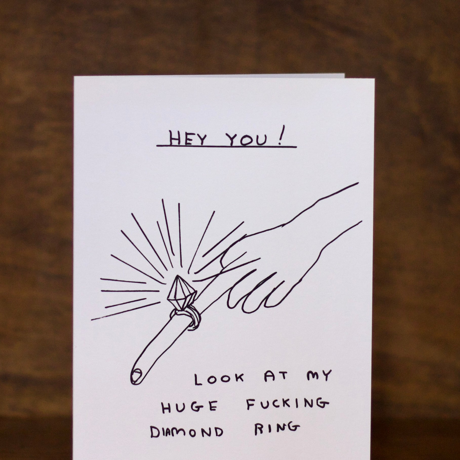 Engagement greeting card with a humorous illustration of a hand holding a diamond ring and text on a wooden background