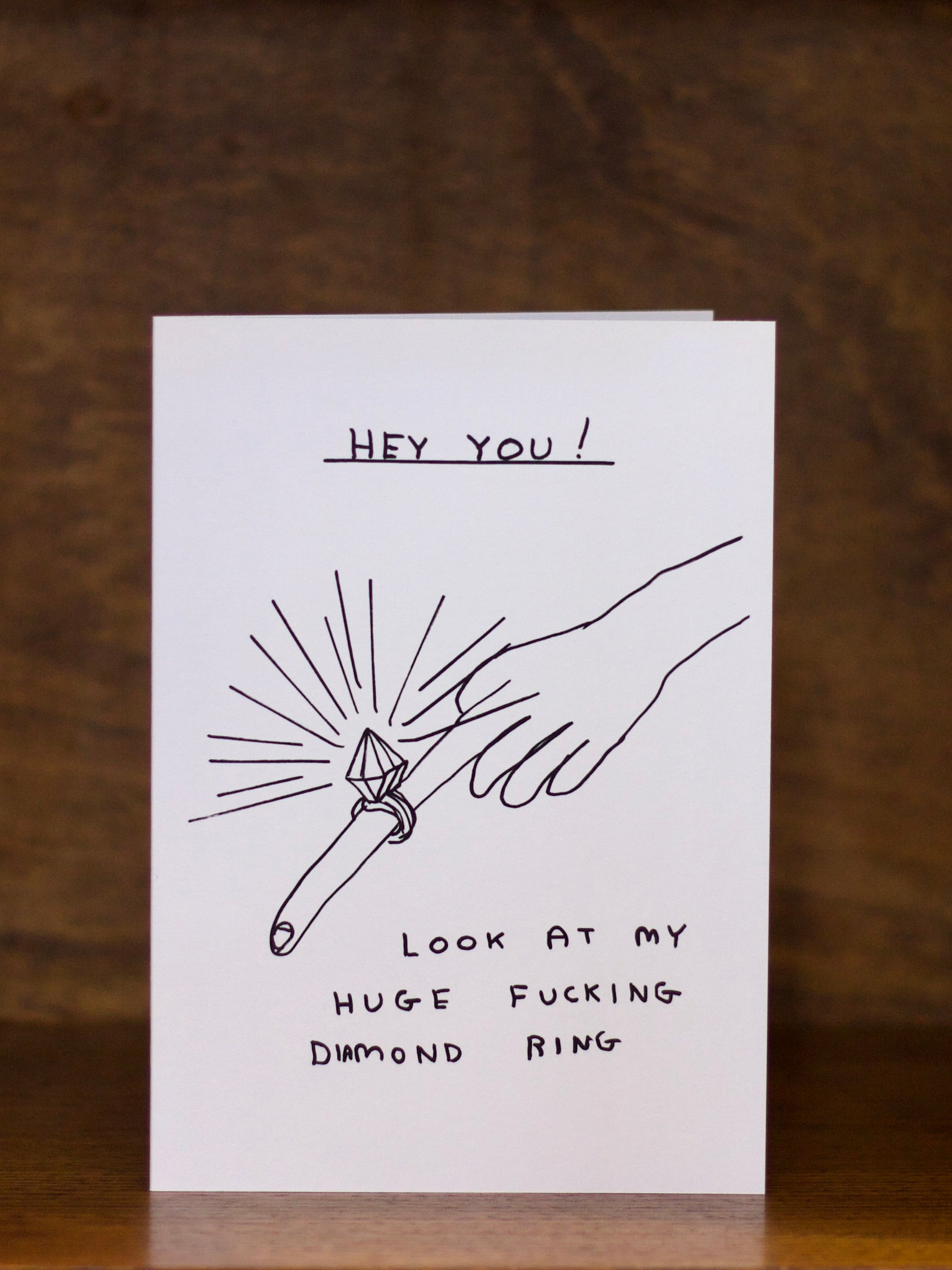 Engagement greeting card with a humorous illustration of a hand holding a diamond ring and text on a wooden background
