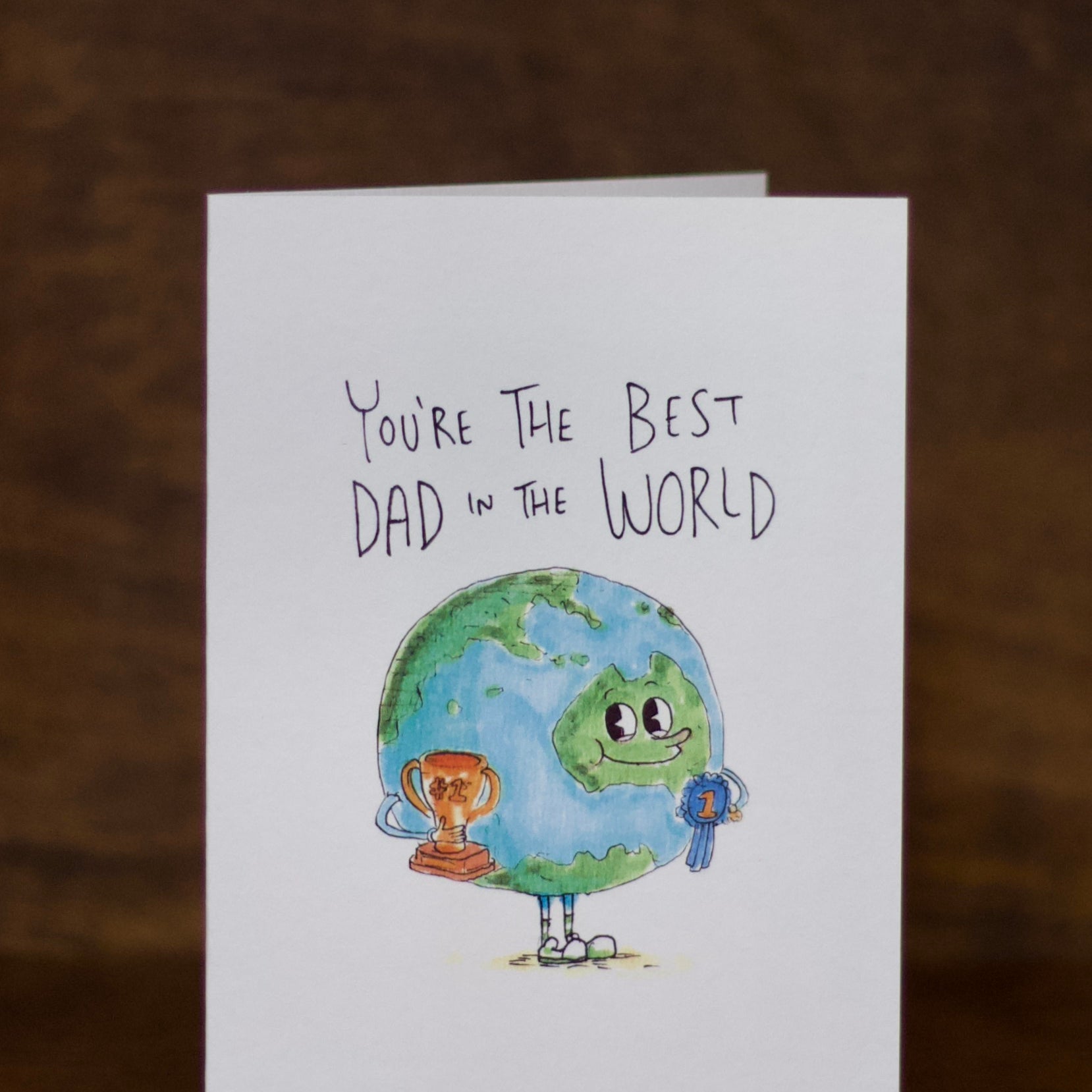 Dad greeting card with illustration of Earth and text 'You're the Best Dad in the World' on a wooden surface.