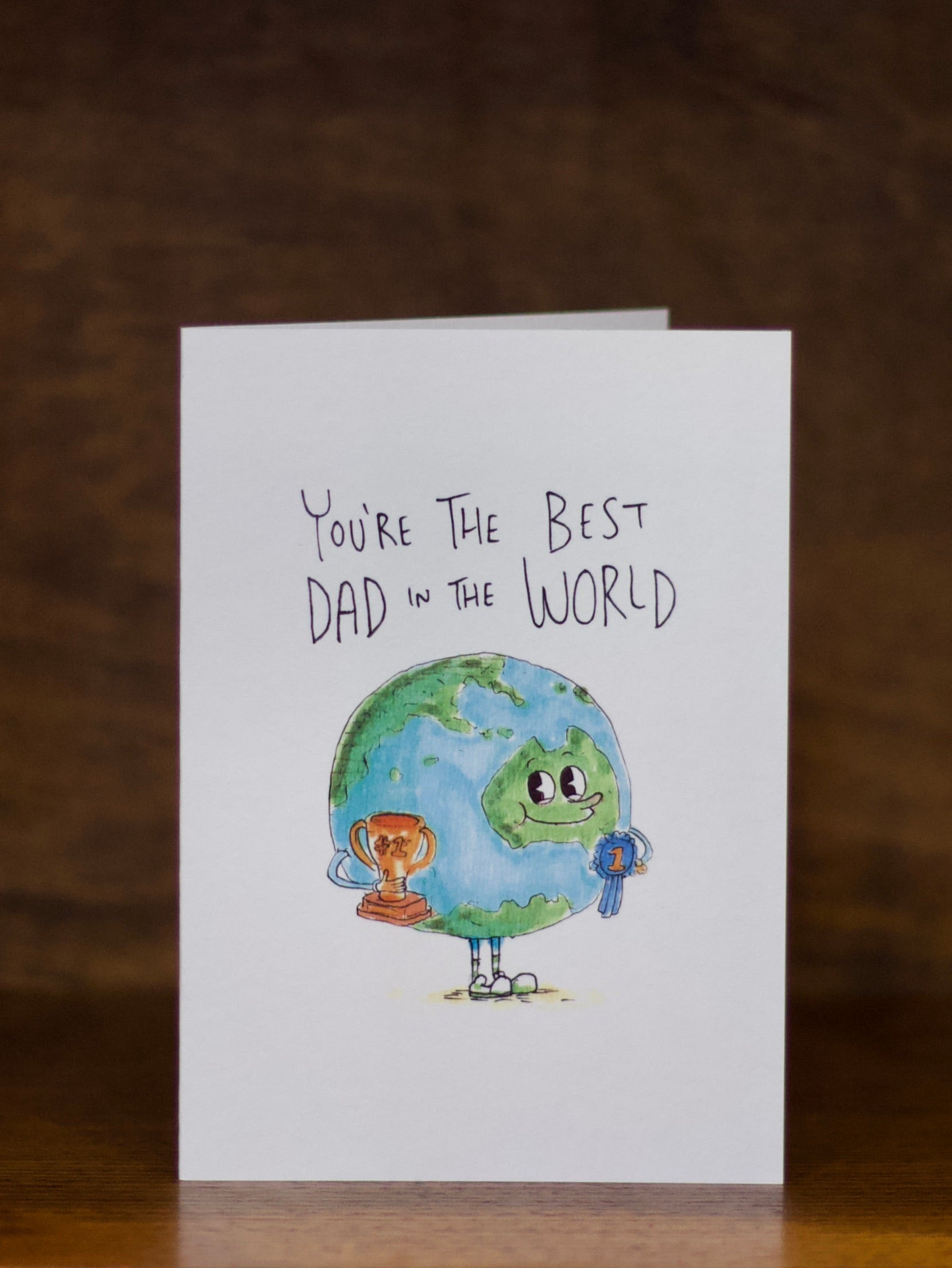 Dad greeting card with illustration of Earth and text 'You're the Best Dad in the World' on a wooden surface.