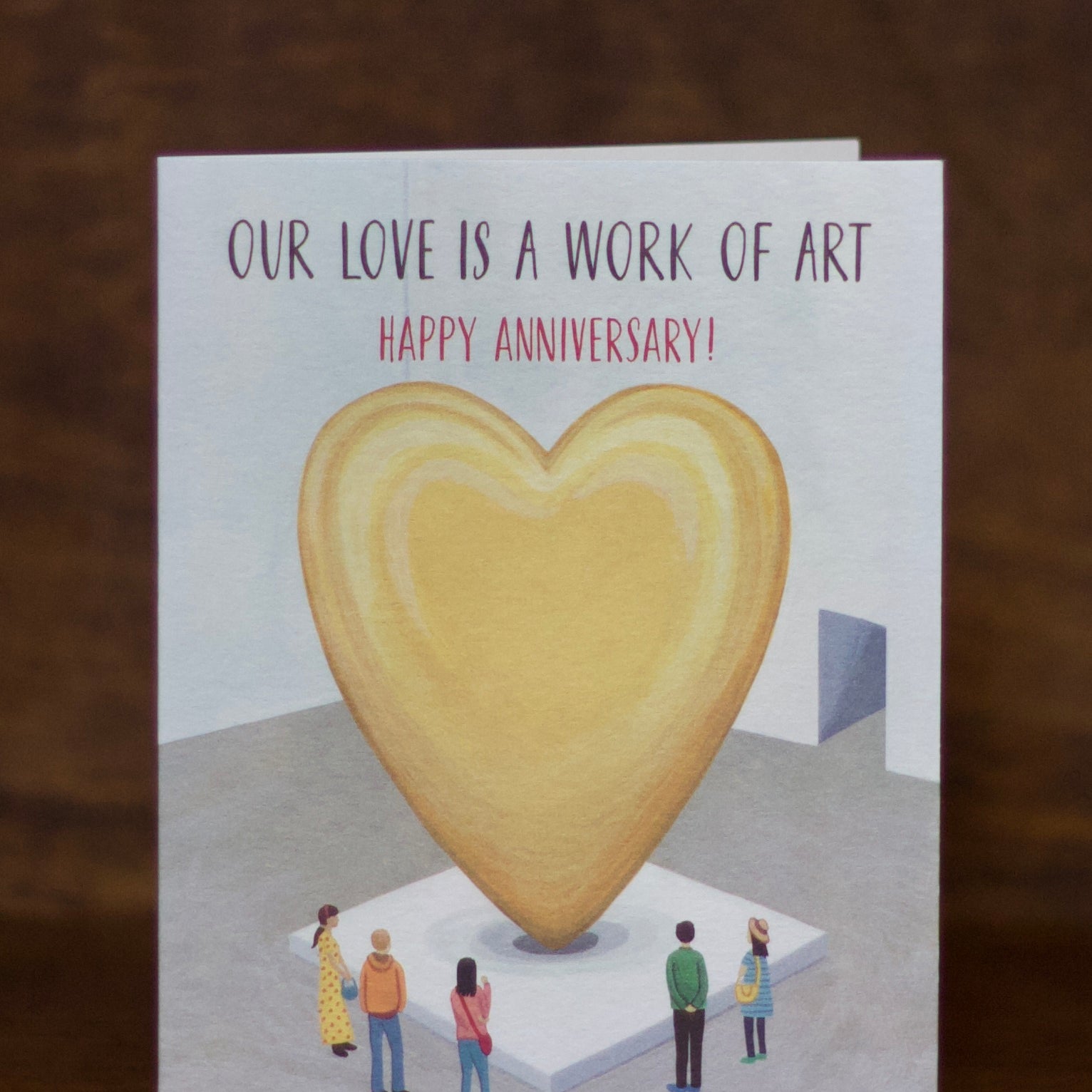 Anniversary greeting card with a heart-shaped sculpture and people admiring it, text 'Our love is a work of art. Happy Anniversary!' on a wooden surface.