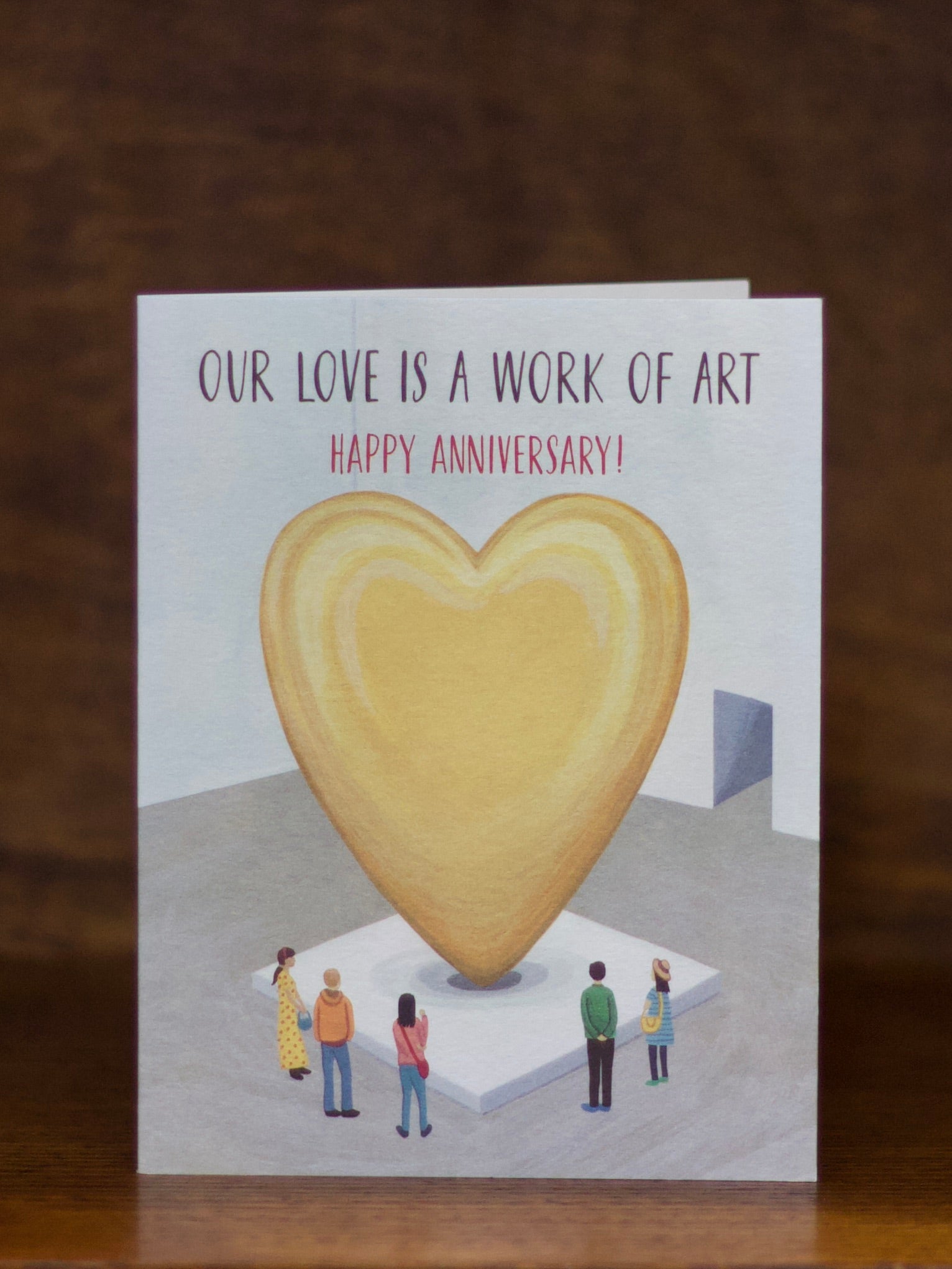 Anniversary greeting card with a heart-shaped sculpture and people admiring it, text 'Our love is a work of art. Happy Anniversary!' on a wooden surface.