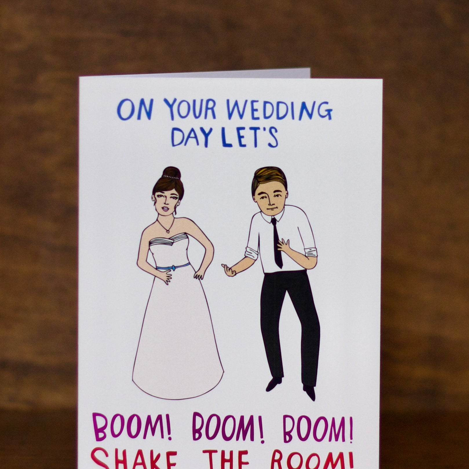 Wedding greeting card with illustrations of a bride and groom and humorous text on a wooden surface.