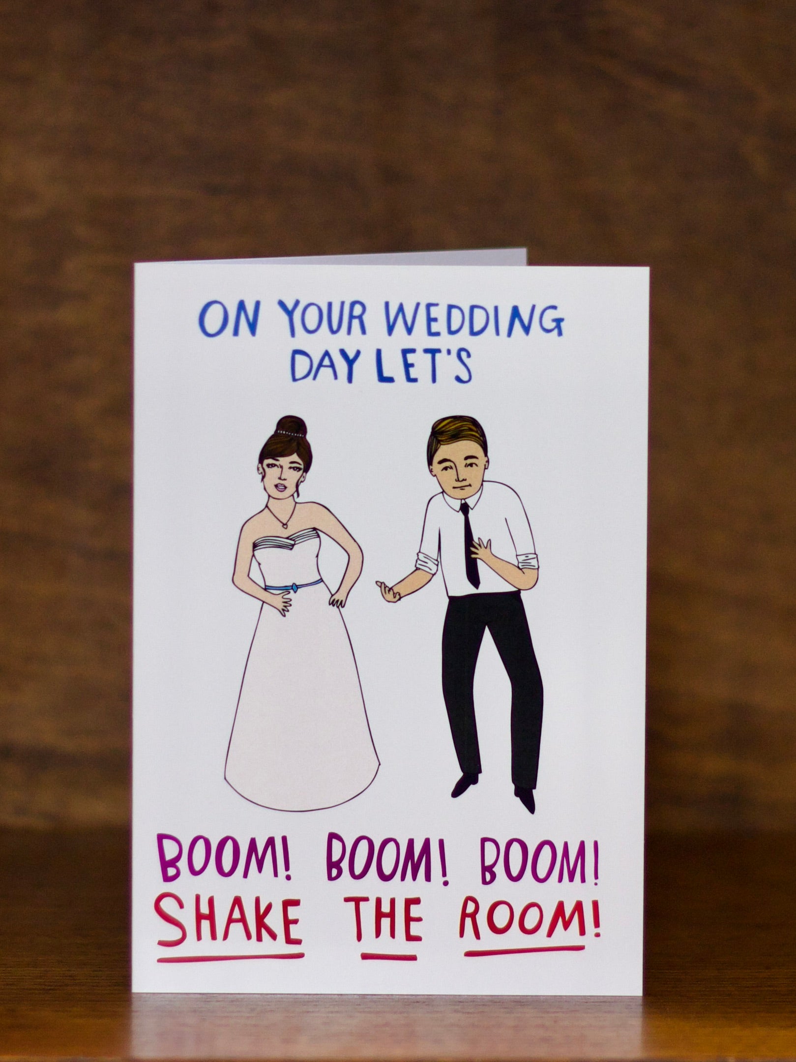 Wedding greeting card with illustrations of a bride and groom and humorous text on a wooden surface.