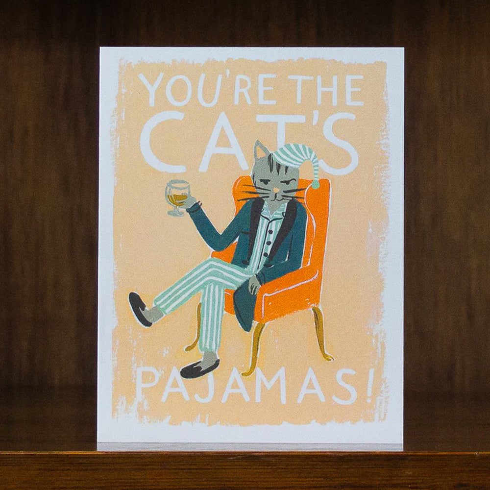 Card with a cat in pajamas holding a glass, text 'You're the cat's pajamas!', on a wooden surface.