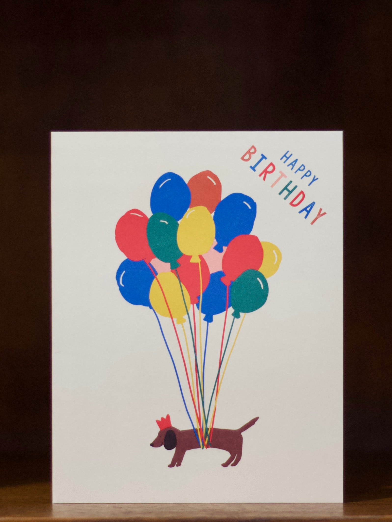 Happy birthday sausage dog wearing a crown floating with lots of balloons.
