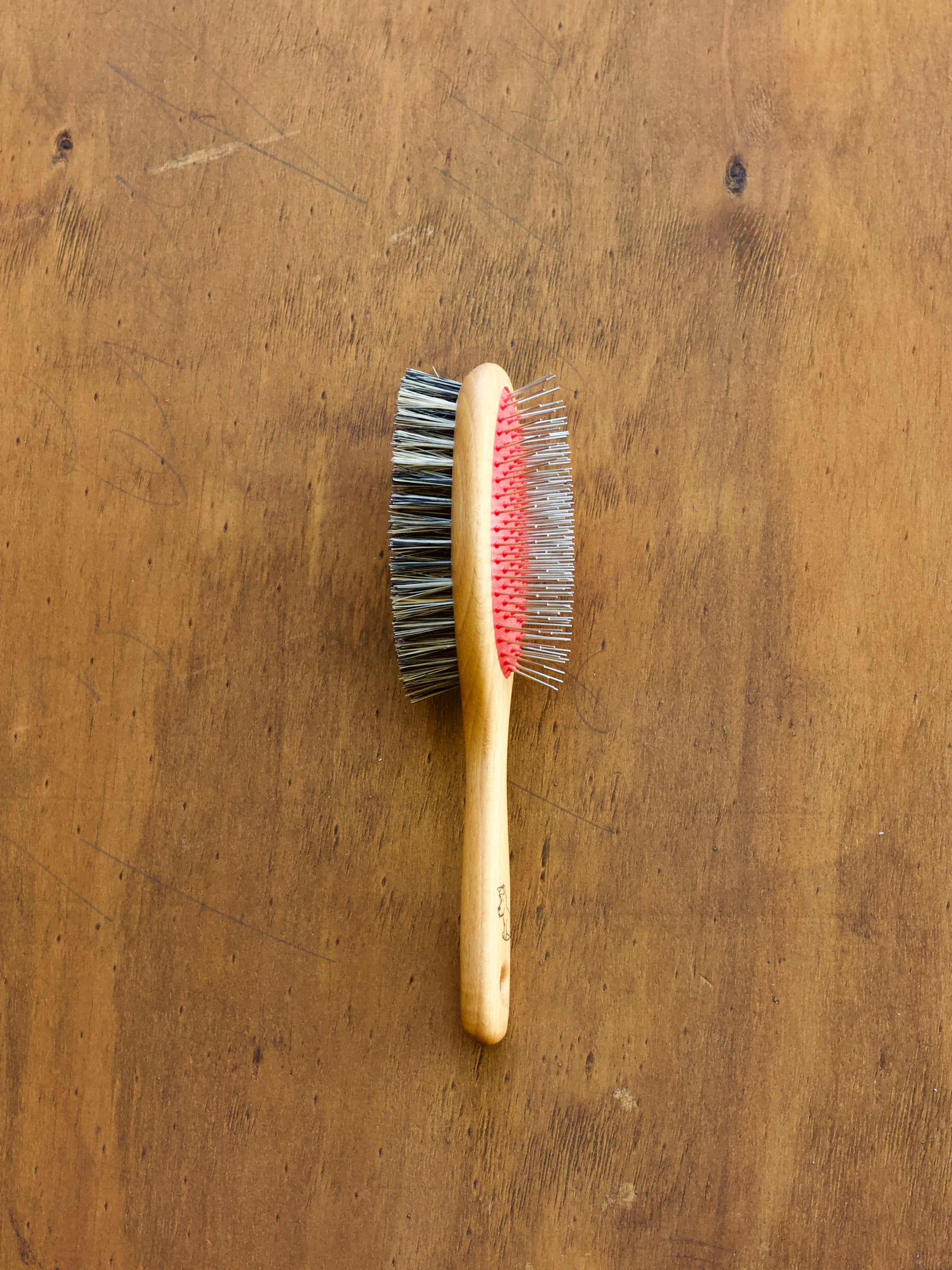 Double-sided pet brush with black bristles on one side and pins on the other, on a wooden surface.