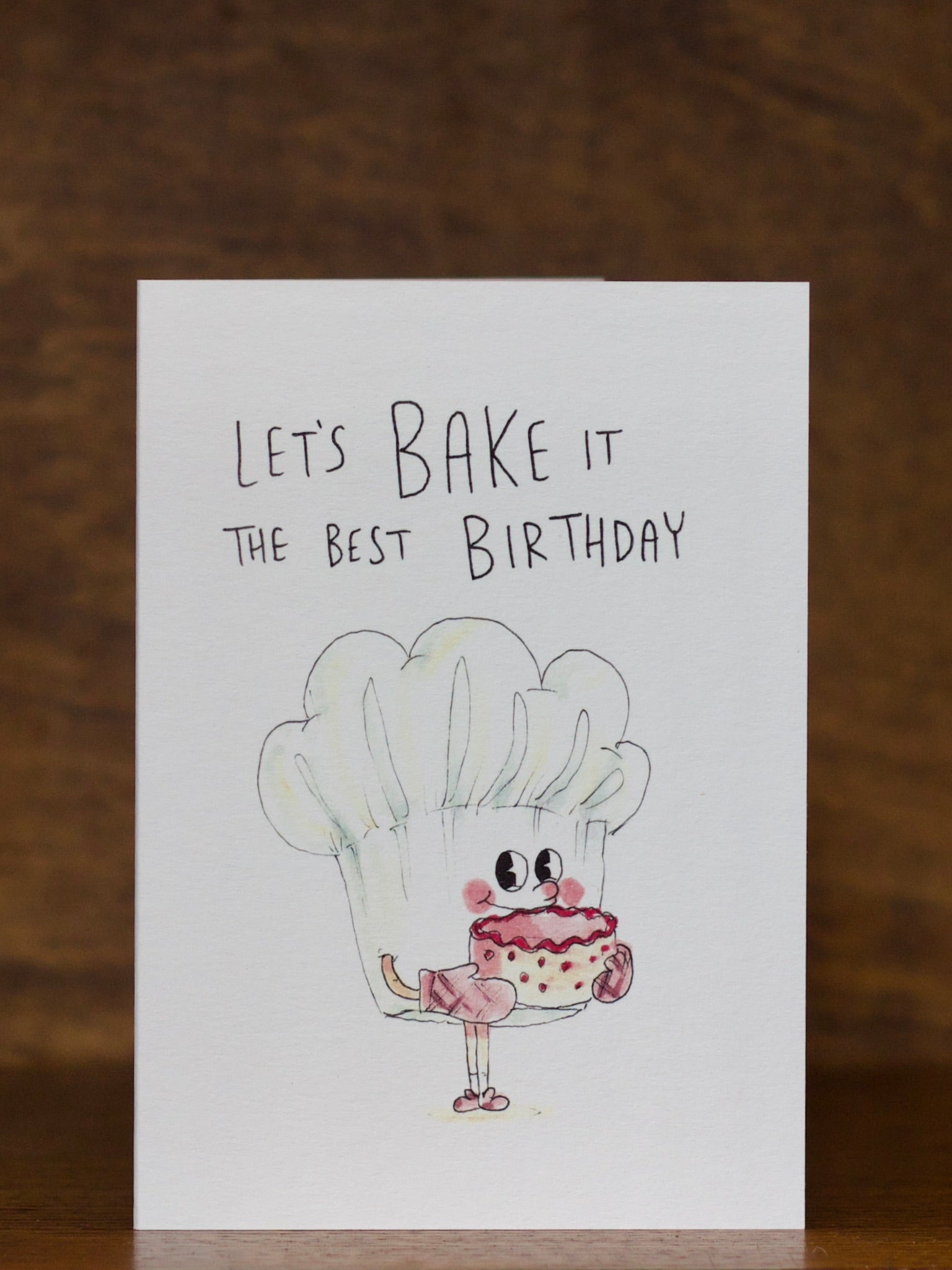 Birthday greeting card by Well Drawn with cartoon character holding a cupcake and text 'Let's Bake it the best Birthday' on a wooden background.