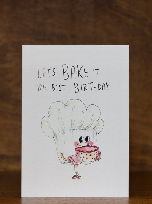 Birthday greeting card by Well Drawn with cartoon character holding a cupcake and text 'Let's Bake it the best Birthday' on a wooden background.