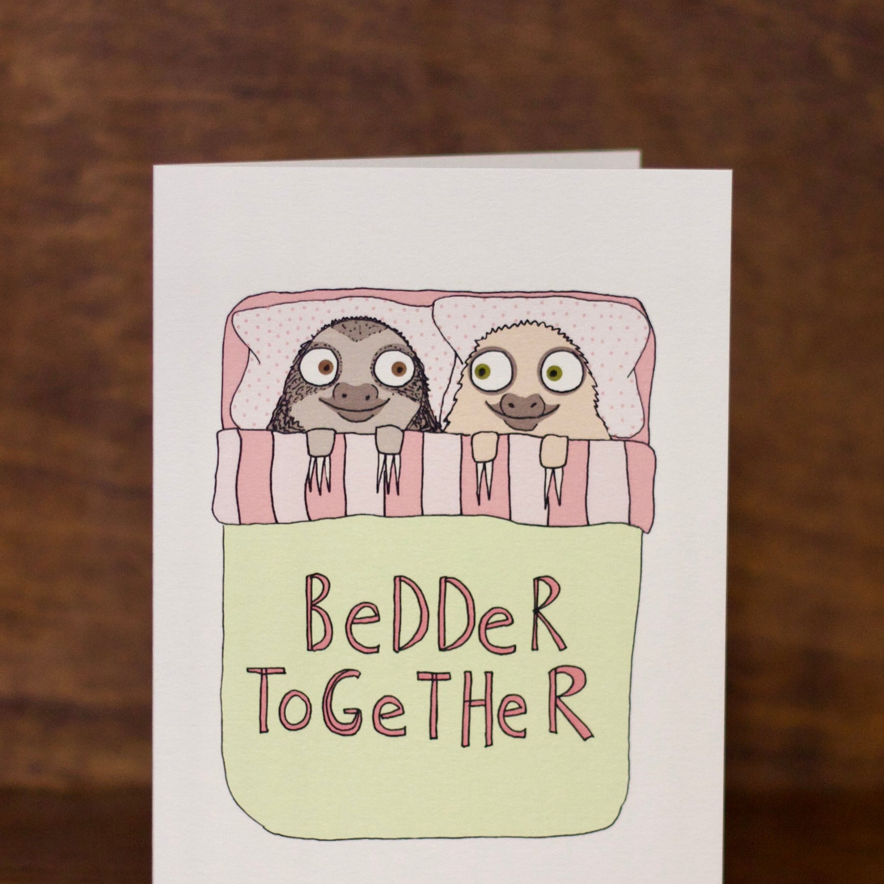 Love & Friendship greeting card with two sloths in bed and 'Bedder Together' text on a wooden background