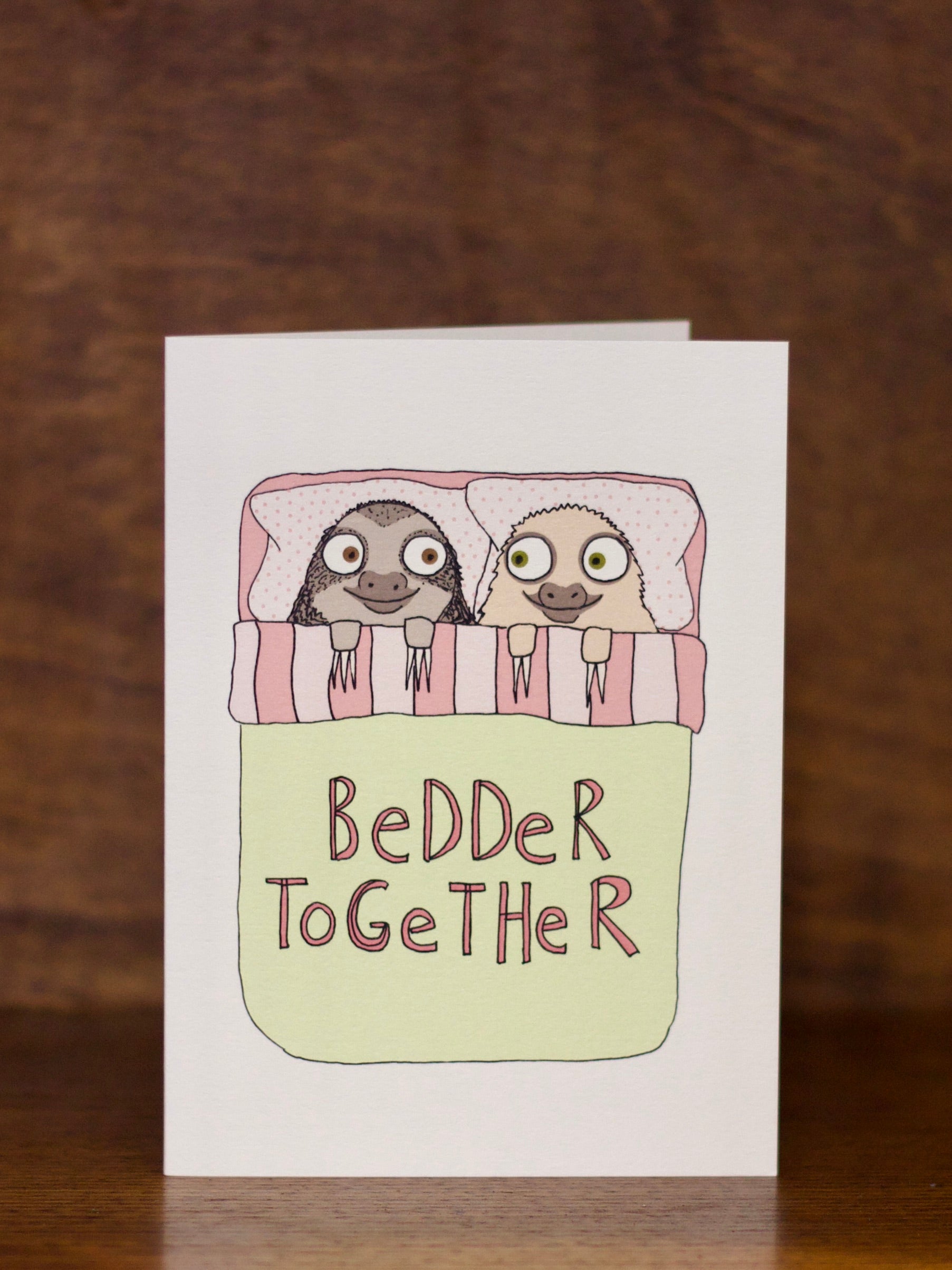 Love & Friendship greeting card with two sloths in bed and 'Bedder Together' text on a wooden background