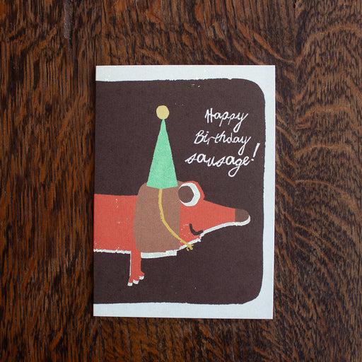 Florence Fry birthday greeting card with a sausage dog illustration and 'Happy Birthday Sausage!' text on a wooden surface.