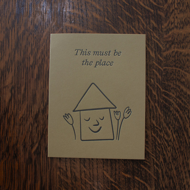 Housewarming greeting card with a house illustration and text 'This must be the place' on a wooden surface
