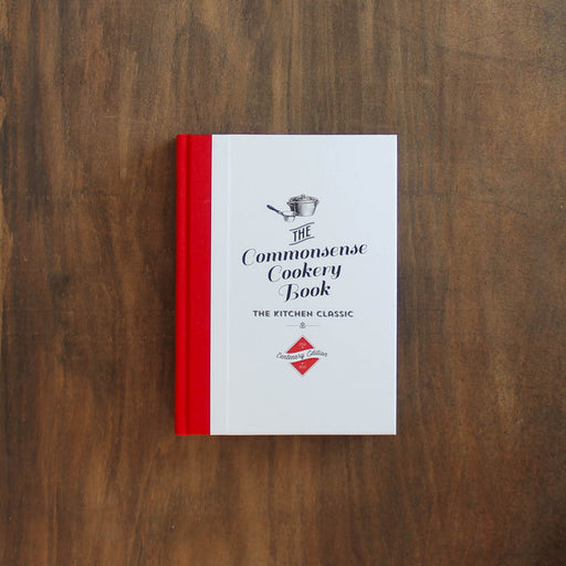 The Commonsense Cookery Book, Volume 2, resting on a dark brown wooden surface. The book features a white cover with black text and illustrations, and a bright red spine.