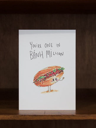 Well Drawn greeting cards with a cartoon banh mi sandwich and text 'You're one in Bánh million' on a wooden surface.